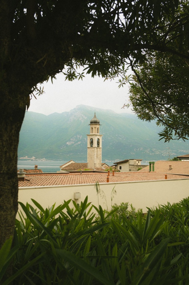 Glockenturm zwischen Olivenzweigen, dahinter Berge am Gardasee